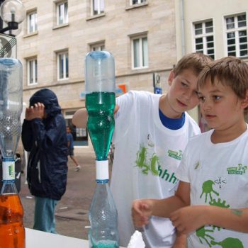 Haus der kleinen Forscher – Kinderprojekte am Stand in der Bayreuther Innenstadt: 2 Jungs rätseln, wie das Wasser von einer Flasche in die andere kommt.