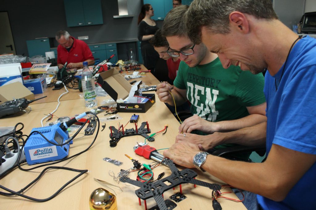 Mehrere Personen bei Elektronikarbeiten im Fablab. Der Tisch ist voll mit Geräten, Lötkolben und Bausätzen