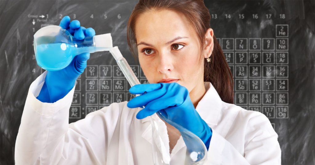 Eine Chemikerin gießt eine blaue Flüssigkeit in ein Reagenzglas. Hinter ihr sind an der Tafel die chemischen Elemente aufgezeichnet