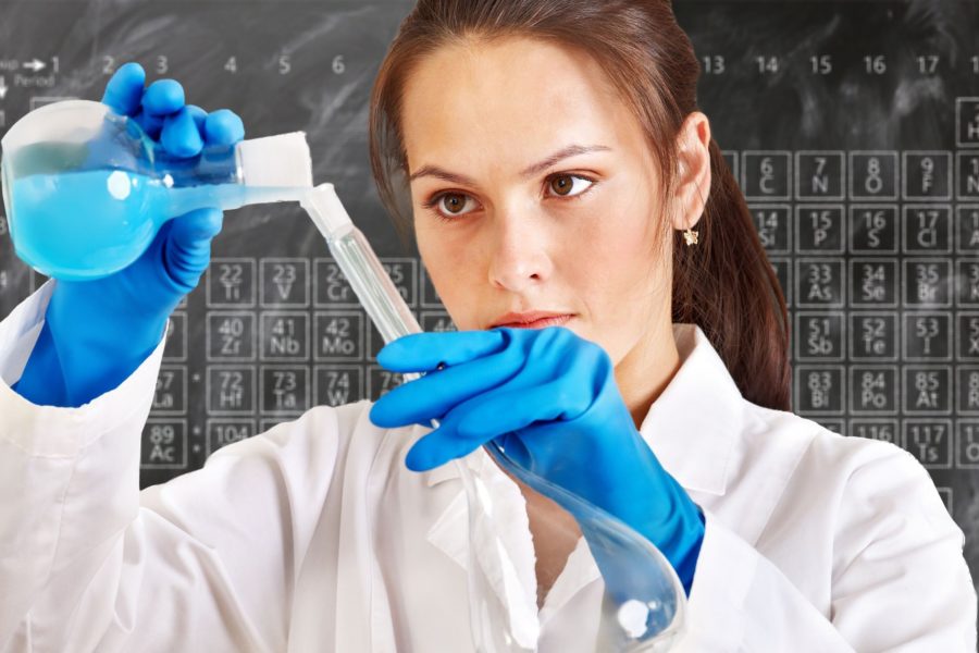 Eine Chemikerin gießt eine blaue Flüssigkeit in ein Reagenzglas. Hinter ihr sind an der Tafel die chemischen Elemente aufgezeichnet