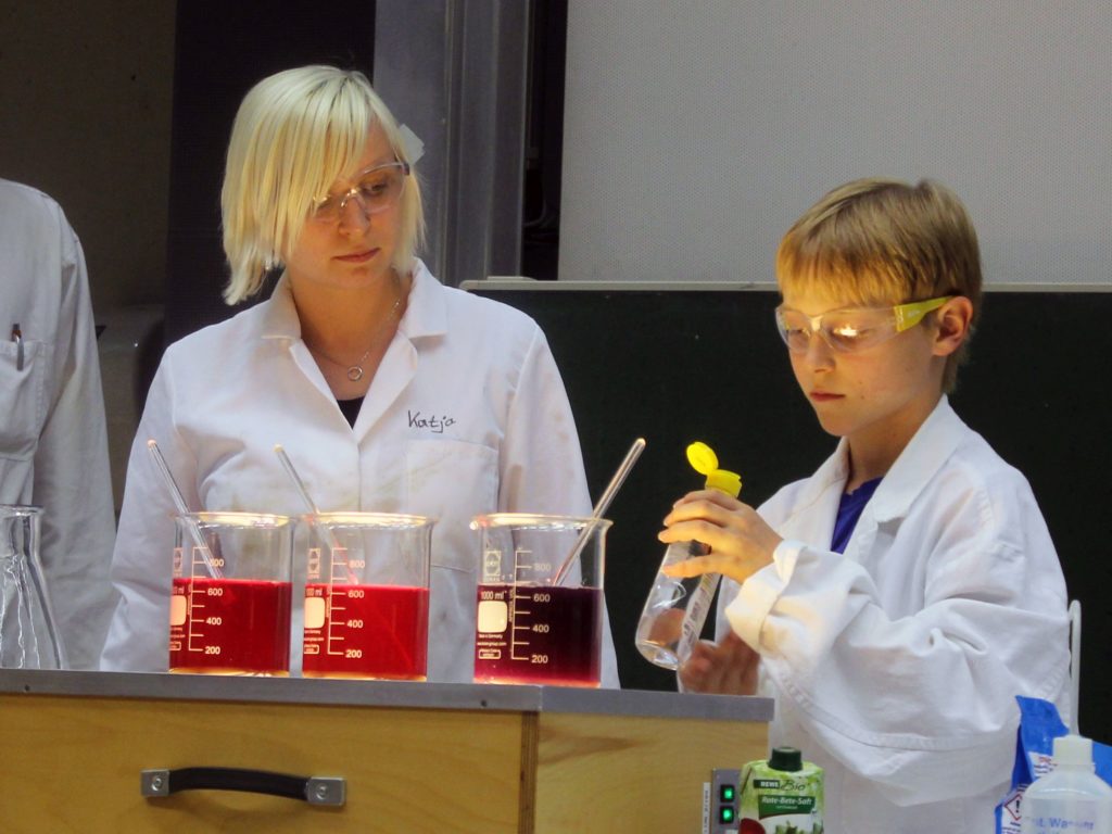 Junge mit Wissenschaftlerin vor verschiedensten Reagenzgläsern.