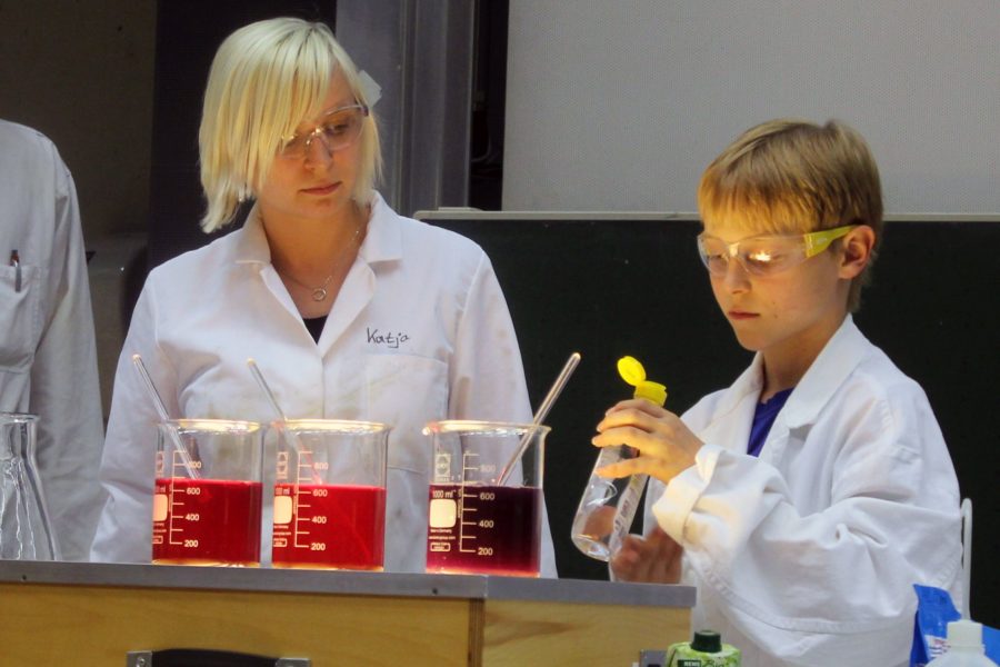 Junge mit Wissenschaftlerin vor verschiedensten Reagenzgläsern.