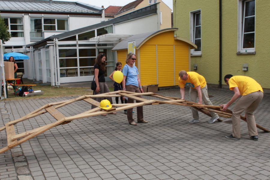 Mehrere Personen bauen eine große Holzbrücke aus holzplanken auf. Stabil genug, um darüber zu gehen.
