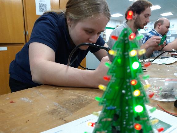 Mehrere Personen beim Elektroniklöten. Im Vordergrund leuchtet ein schon fertiger kleiner Weihnachtsbaum