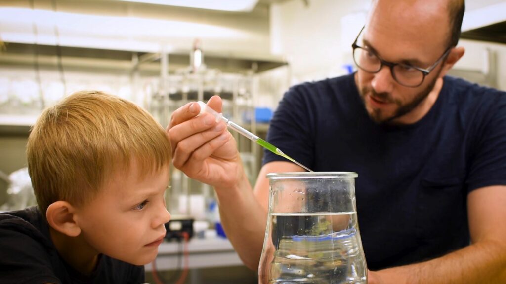 Ein Mann tropft mit einer Pipette eine Flüssigkeit in ein Glas. Ein Junge beobachtet genau, was passiert.