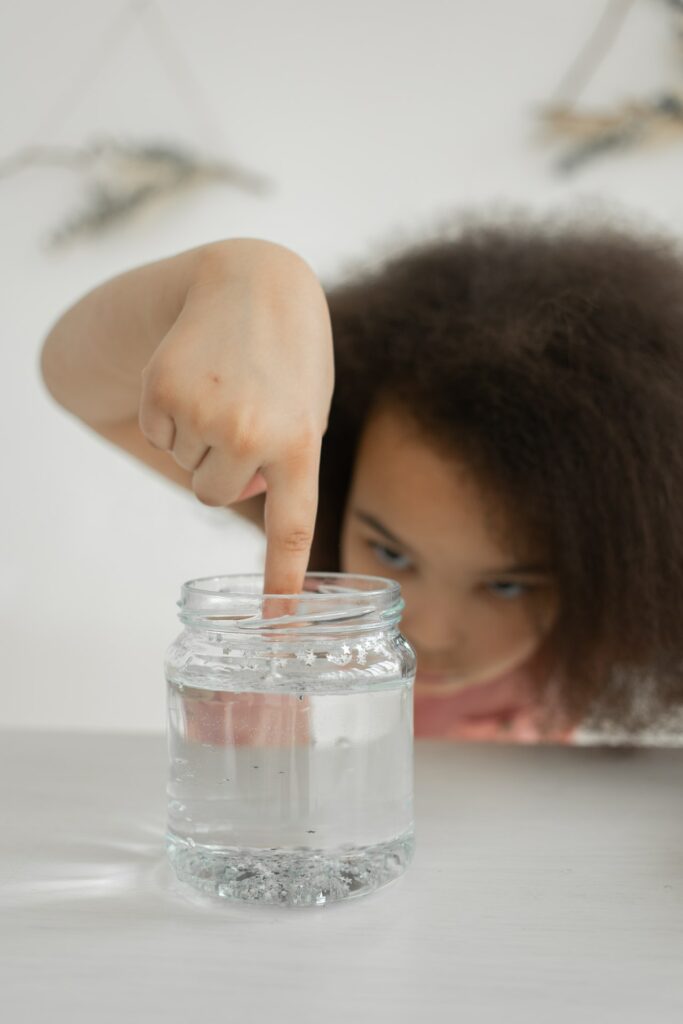 Ein Mädchen steckt vorsichtig seinen Finger in ein Glas mit durchsichtiger Flüssigkeit.