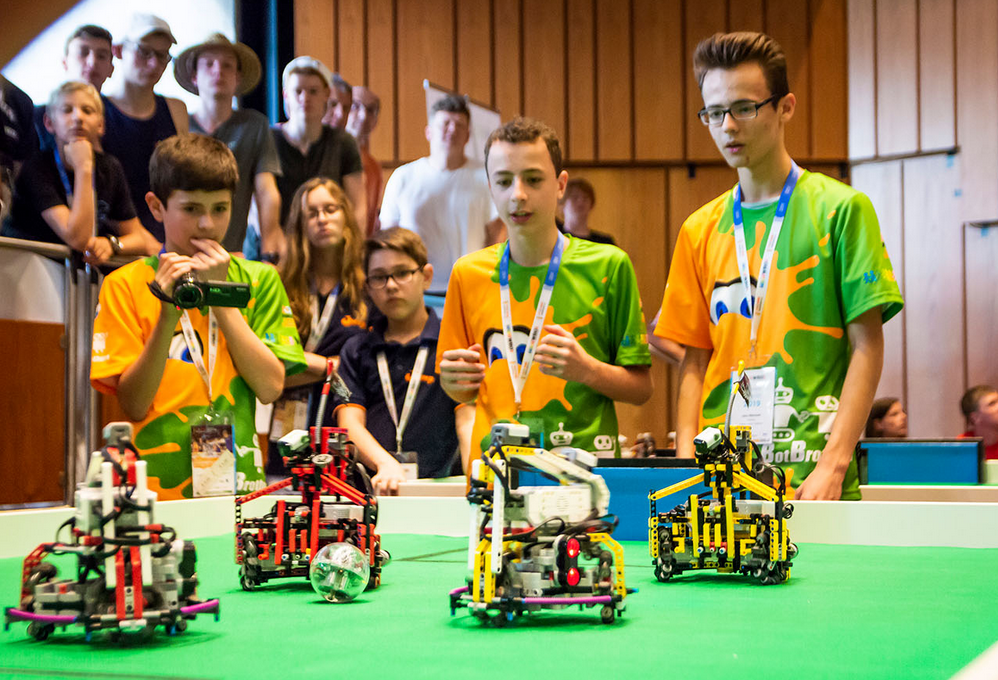 Jugendliche spielen mit vier Robotern Fußball, ein Jugendlicher filmt das Spiel. Im Hintergrund sieht man zahlreiche Zuschauer.