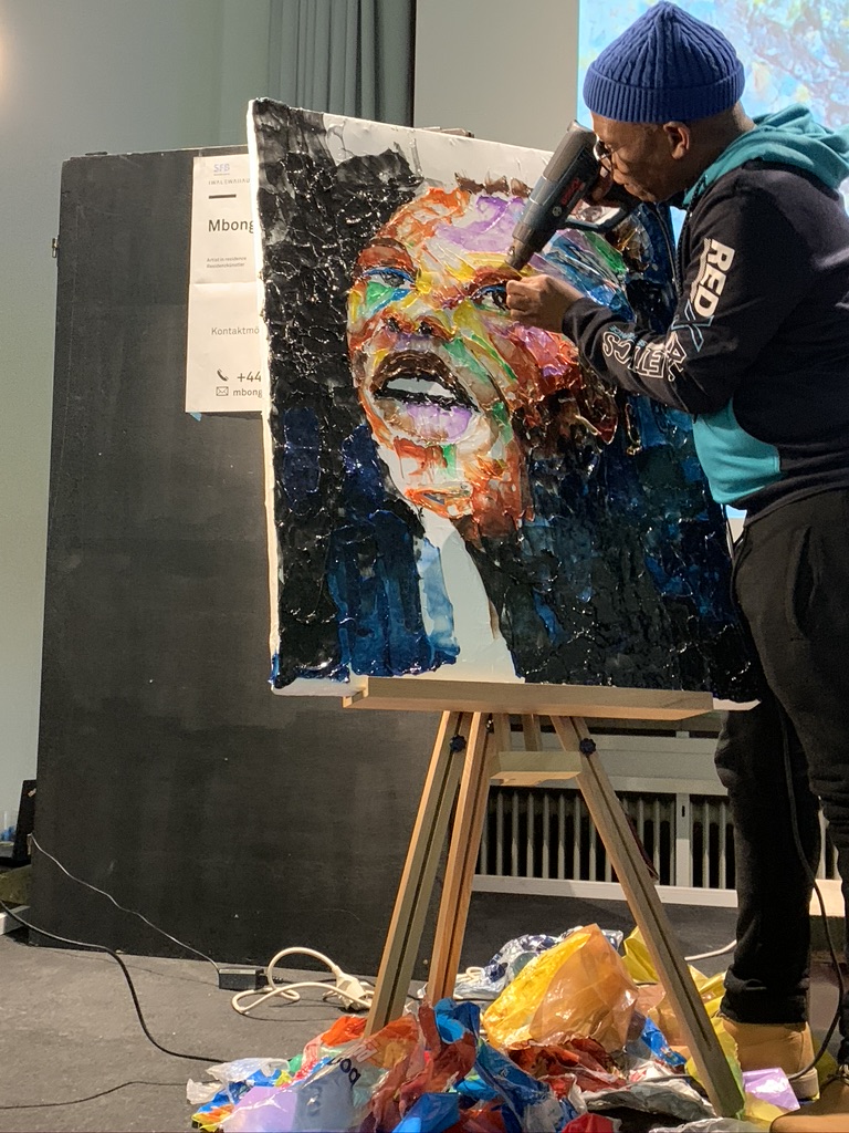Ein Kunstschaffender bearbeitet mit einem Föhn ein Gemälde, das ein Gesicht zeigt. Am Boden liegen zahlreiche bunte Plastiktüten